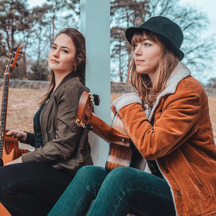 Portaitfoto Sophie Chassée und Karlijn Langendijk. Sie sitzen draußen unter einem Vordach und haben beide Gitarren in der Hand.