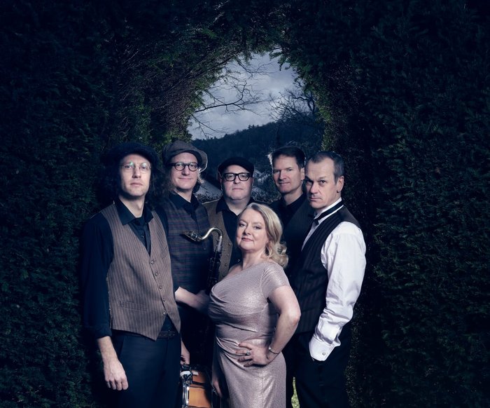 Die Musiker:innen von "Youkali" stehen vor einem pflanzenbewachsenen Torbogen, das Foto hat eine dunkle, aber keine düstere Atmosphäre