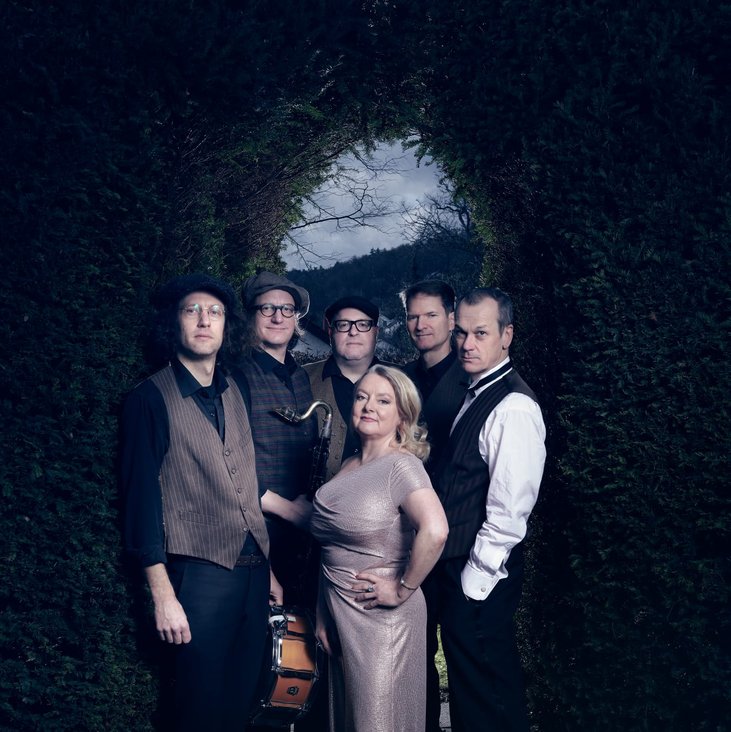 Die Musiker:innen von "Youkali" stehen vor einem pflanzenbewachsenen Torbogen, das Foto hat eine dunkle, aber keine düstere Atmosphäre