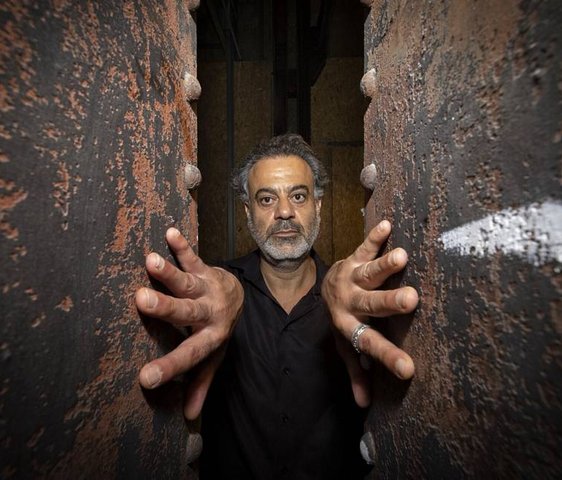 Porträt eines Mannes zwischen zwei rauen Wänden, Hände an den Flächen abgestützt, direkter Blick.