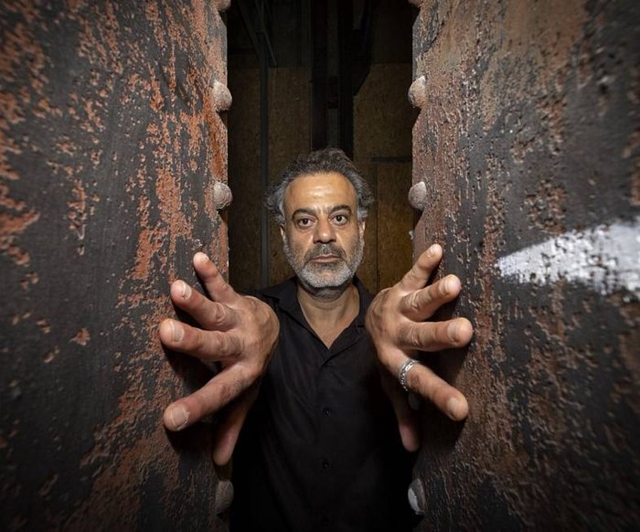 Porträt eines Mannes zwischen zwei rauen Wänden, Hände an den Flächen abgestützt, direkter Blick.