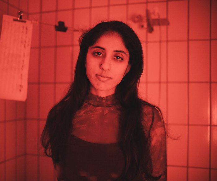 Die Künstlerin steht in einem rot beleuchtetem gefließtem Raum und schaut in die Kamera.