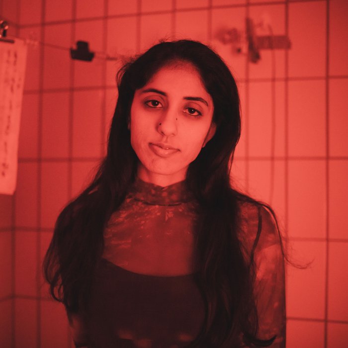 Die Künstlerin steht in einem rot beleuchtetem gefließtem Raum und schaut in die Kamera.