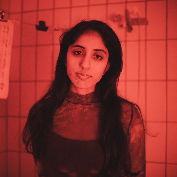 Die Künstlerin steht in einem rot beleuchtetem gefließtem Raum und schaut in die Kamera.