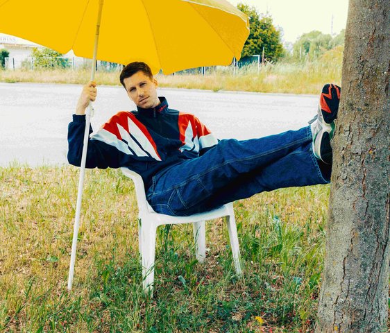 Der Künstler sitzt auf einem Plastikstuhl auf einer Wiese. Seine Beine sind ausgestreckt an einem Baumstamm abgestützt und er hält sich an einem gelben Sonnenschirm fest.