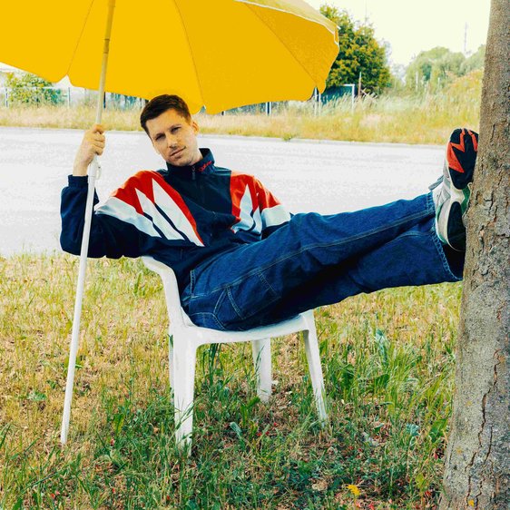 Der Künstler sitzt auf einem Plastikstuhl auf einer Wiese. Seine Beine sind ausgestreckt an einem Baumstamm abgestützt und er hält sich an einem gelben Sonnenschirm fest.