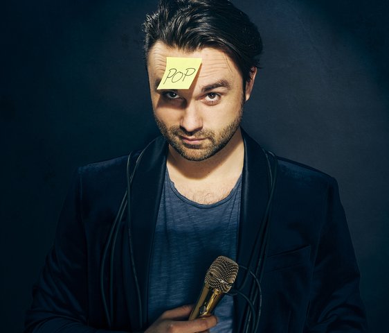 Ein Mann blickt in die Kamera, in der Hand hält er ein Mikrofon, auf der Stirn klebt ein Post-It-Zettel mit der handschriftlichen Aufschrift "Pop"