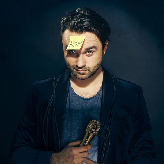 Ein Mann blickt in die Kamera, in der Hand hält er ein Mikrofon, auf der Stirn klebt ein Post-It-Zettel mit der handschriftlichen Aufschrift "Pop"