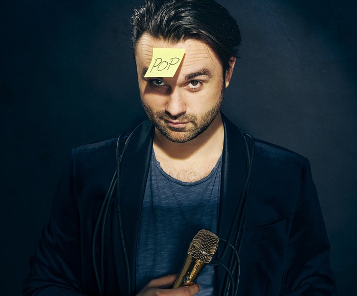 Ein Mann blickt in die Kamera, in der Hand hält er ein Mikrofon, auf der Stirn klebt ein Post-It-Zettel mit der handschriftlichen Aufschrift "Pop"