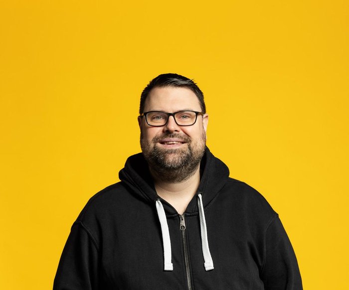 Philipp Uckel lacht vor gelben Hintergrund in einem lässigen Outfit in die Kamera