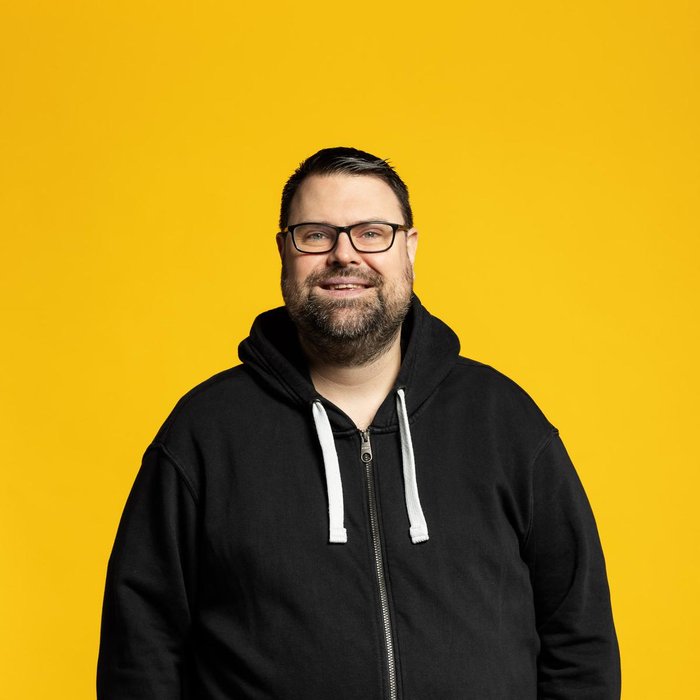 Philipp Uckel lacht vor gelben Hintergrund in einem lässigen Outfit in die Kamera