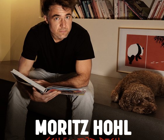 Moritz Hohl blickt in die Kamera, ein geöffnetes Buch auf dem Schoß, ein Hund liegt neben ihm.