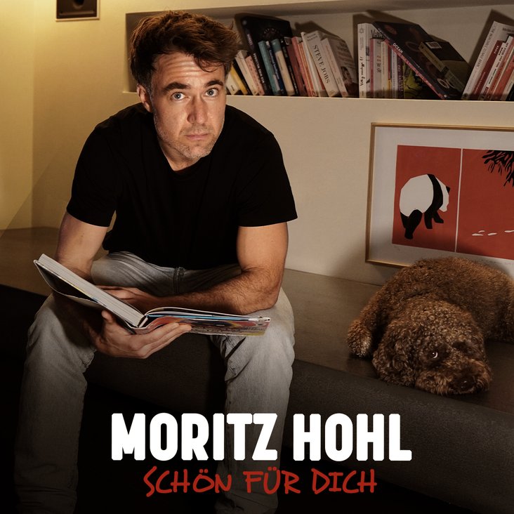 Moritz Hohl blickt in die Kamera, ein geöffnetes Buch auf dem Schoß, ein Hund liegt neben ihm.