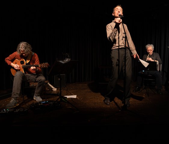 Ein Mann steht auf der Bühne und spricht, neben ihm eine Band aus zwei weiteren Männern.