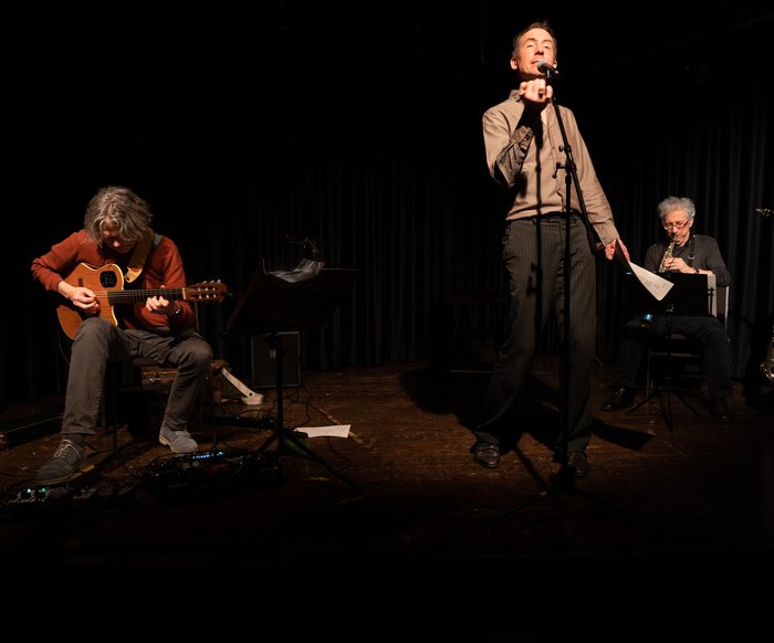 Ein Mann steht auf der Bühne und spricht, neben ihm eine Band aus zwei weiteren Männern.