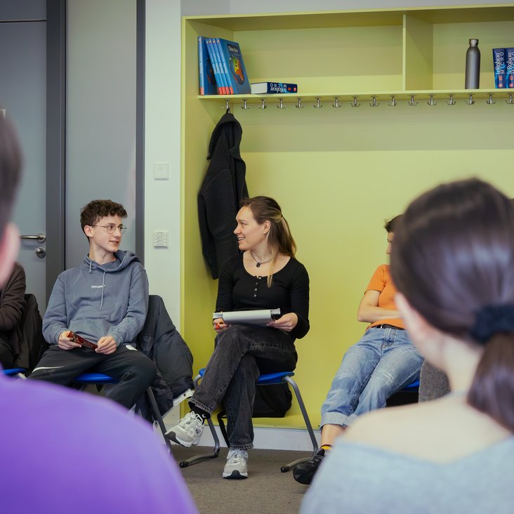 Eine Theaterpädagogin sitzt im Stuhlkreis mit einer Klasse und bespricht das Stück nach.