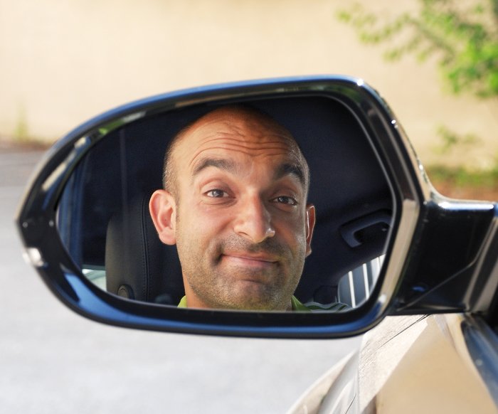 Django Asül lächelt. Man sieht sein Spiegelbild im Rückspiegel eines Autos.