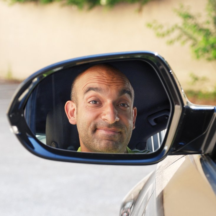 Django Asül lächelt. Man sieht sein Spiegelbild im Rückspiegel eines Autos.