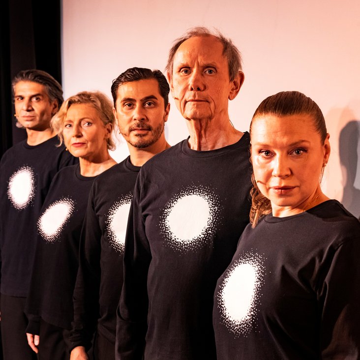 Die Schauspieler:innen von "And now Hanau" auf der Bühne. Sie tragen alle schwarze Kleidung mit einem vollflächig weißen Kreis auf der Brust.