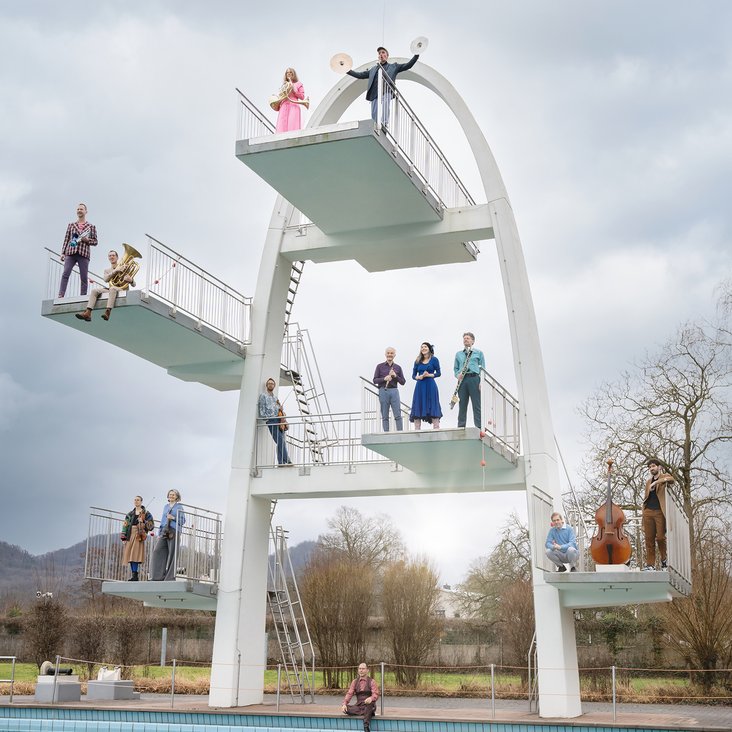 Die Musiker sind auf die unterschiedlichen Plattformen eines Sprungturms im Schwimmbad verteilt und tragen bunte Kleidung.