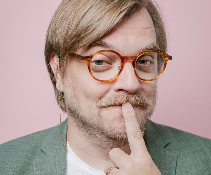 Auf dem Foto ist Nilz Bokelberger mit einer Brille zu sehen. Er legt den Zeigefinger an seinen Mund. Er trägt ein weißes Shirt mit lila Aufschrift und ein graugrünes Sakko.