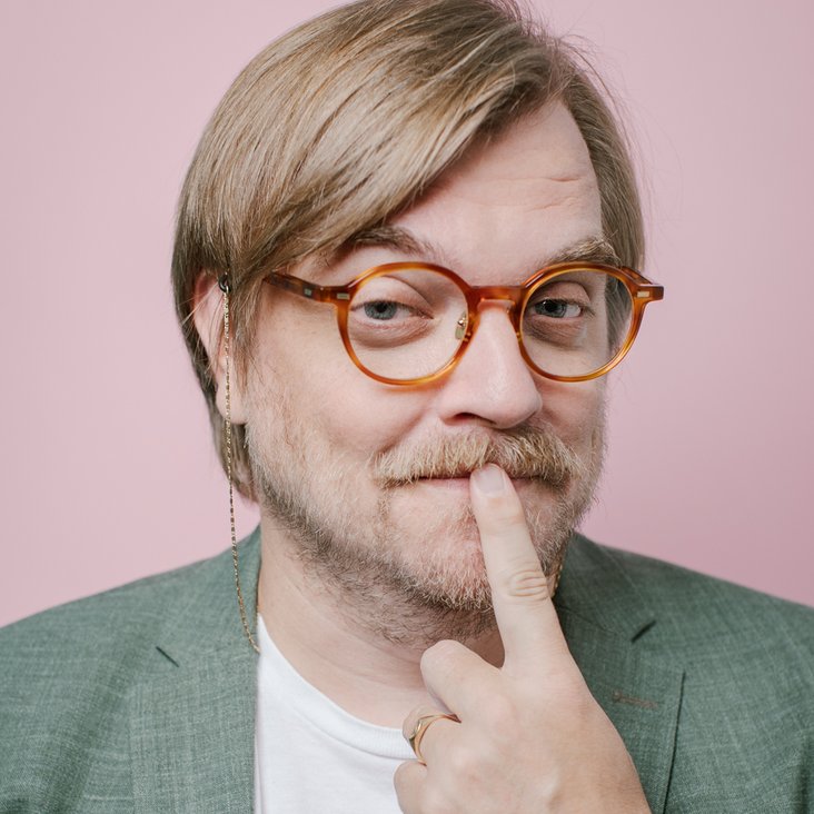 Auf dem Foto ist Nilz Bokelberger mit einer Brille zu sehen. Er legt den Zeigefinger an seinen Mund. Er trägt ein weißes Shirt mit lila Aufschrift und ein graugrünes Sakko.