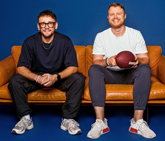 Die zwei Podcaster sitzen nebeneinander auf einer Couch und schauen lächelnd in die Kamera.