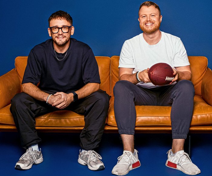 Die zwei Podcaster sitzen nebeneinander auf einer Couch und schauen lächelnd in die Kamera.