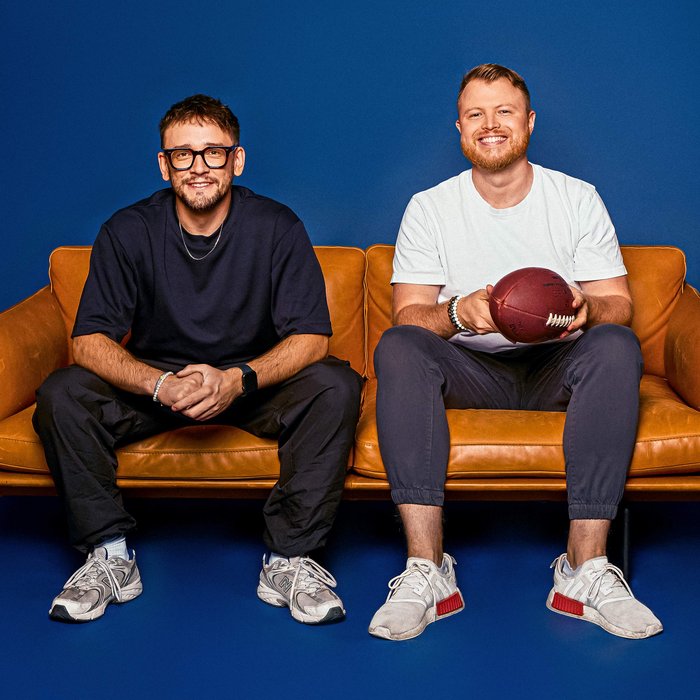 Die zwei Podcaster sitzen nebeneinander auf einer Couch und schauen lächelnd in die Kamera.