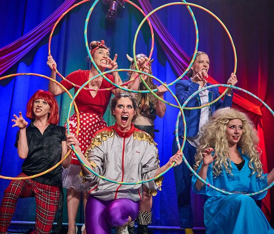 Die Künstlerinnen der Barbaren Barbies in Kostüm auf der Bühne. Jede hält einen Hula Hoop Reifen in der Hand, durch den sie blickt.