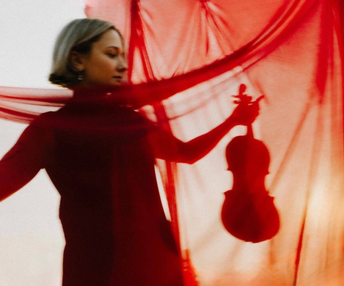 Performerin mit Violine hinter rotem Stoff, das Instrument und Körperhaltung zeichnen sich als Silhouette ab.