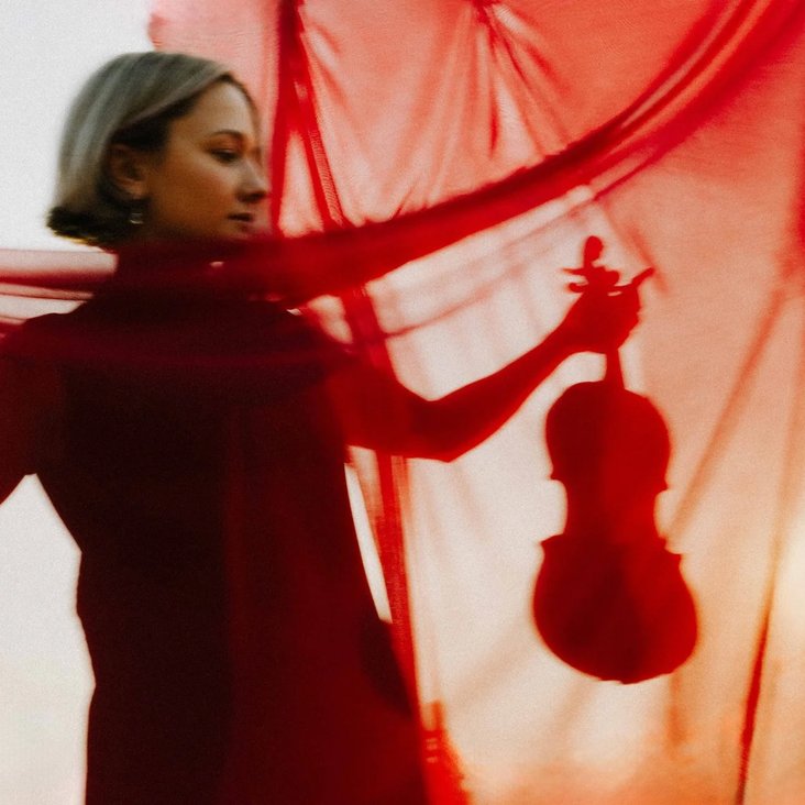 Performerin mit Violine hinter rotem Stoff, das Instrument und Körperhaltung zeichnen sich als Silhouette ab.