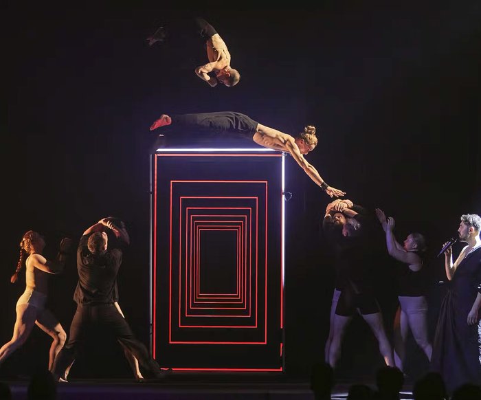 Verschiedene Artist*innen auf der Bühne. Das Bühnenbild ist eher düster & futuristisch, ebenso die Kostüme.