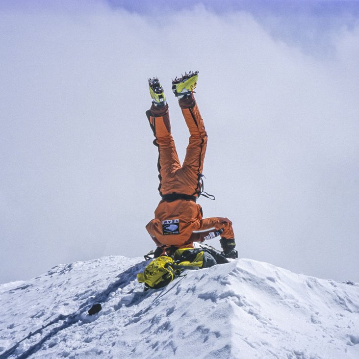 Ein Mensch in orangefarbenem Schneenanzug und Schuhen mit Spikes ist zu sehen. Er macht auf einem schneebdeckten Berggipfel einen Kopfstand.