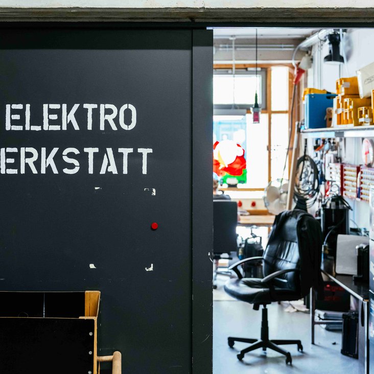 Tür mit Schriftzug „ELEKTRO WERKSTATT“, Werkraum dahinter.