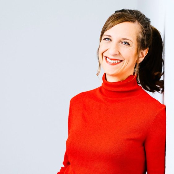 Sarah Hakenberg trägt blaue Jeans und einen roten Rollkragenpullover. Sie hat die Hände in den Taschen und lehnt an eine Wand und lächelt in die Kamera.