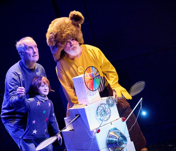 Bühnenszene aus einem Musiktheaterstück: zwei Performer und eine Figur im Bärenkostüm stehen beleuchtet auf einer Theaterbühne.