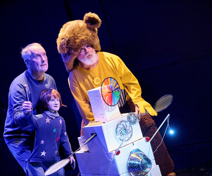 Bühnenszene aus einem Musiktheaterstück: zwei Performer und eine Figur im Bärenkostüm stehen beleuchtet auf einer Theaterbühne.