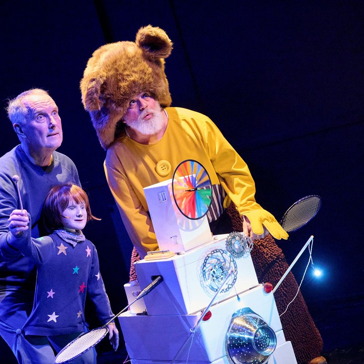 Bühnenszene aus einem Musiktheaterstück: zwei Performer und eine Figur im Bärenkostüm stehen beleuchtet auf einer Theaterbühne.