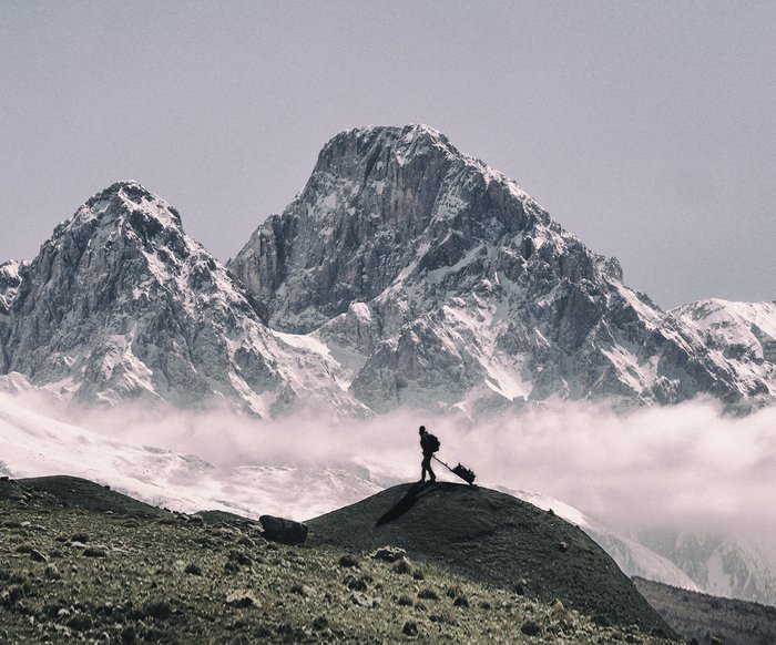 Eine Person geht zu Fuß vor einem großen, schneebedeckten Bergmassiv und zieht einen Rollkoffer oder etwas ähnliches hinter sich her