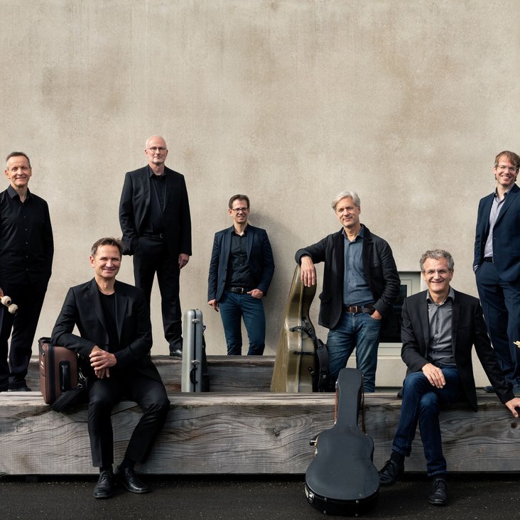 Gruppenporträt eines Ensembles vor einer steinernen Wand, Musikerinnen und Musiker in formeller Kleidung.