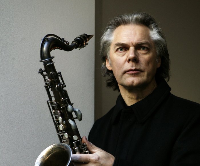 Jan Garbarek mit Saxophoh in der Hand vor grauem Hintergrund.