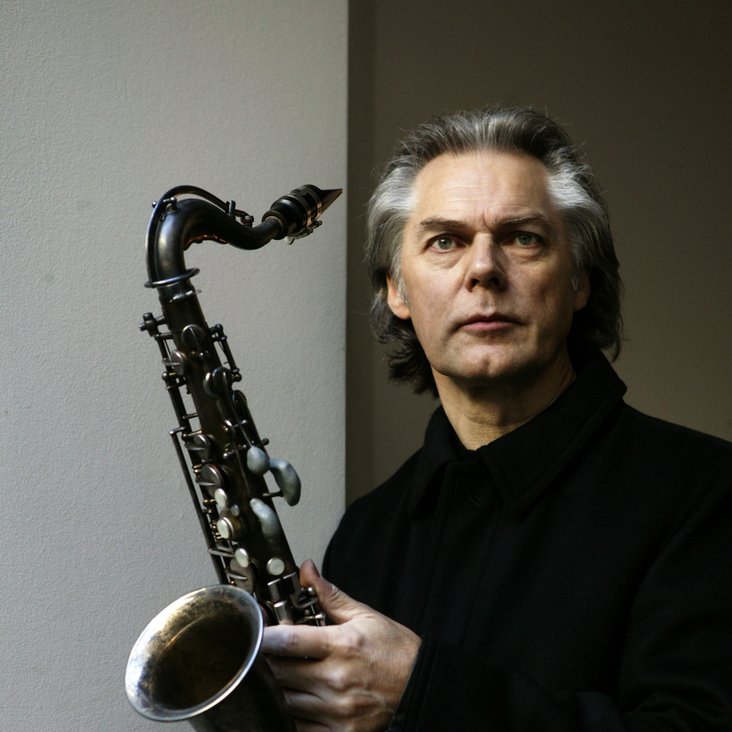 Jan Garbarek mit Saxophoh in der Hand vor grauem Hintergrund.
