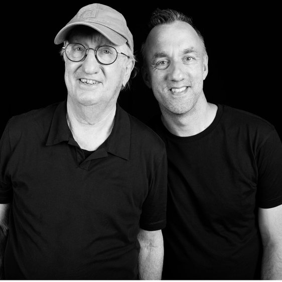 Ein schwarz-weiß Foto von Jan-Uwe Rogge und Matthias Jung. Die beiden stehen an einem Stehtisch und blicken in Richtung der Kamera.