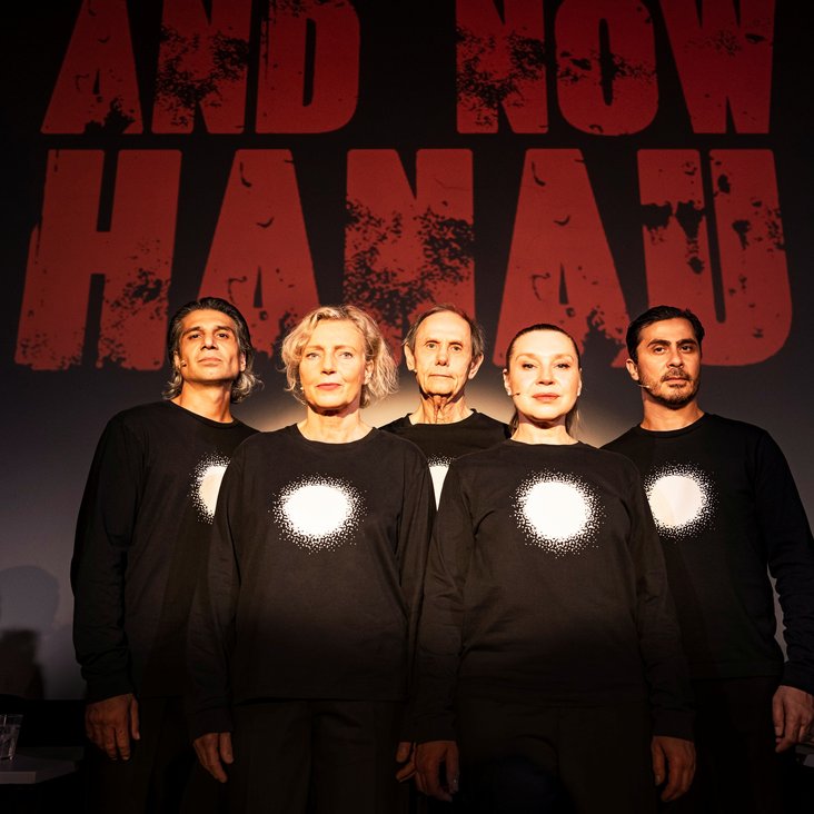 Die Schauspieler:innen von "And now Hanau" auf der Bühne. Sie tragen alle schwarze Kleidung mit einem vollflächig weißen Kreis auf der Brust.