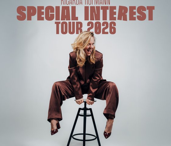 Ricardo Hoffmann in einem braunen glänzenden Jumpsuit auf einem Barhocker. Sie drückt sich mit den Händen auf der Sitzfläche hoch.