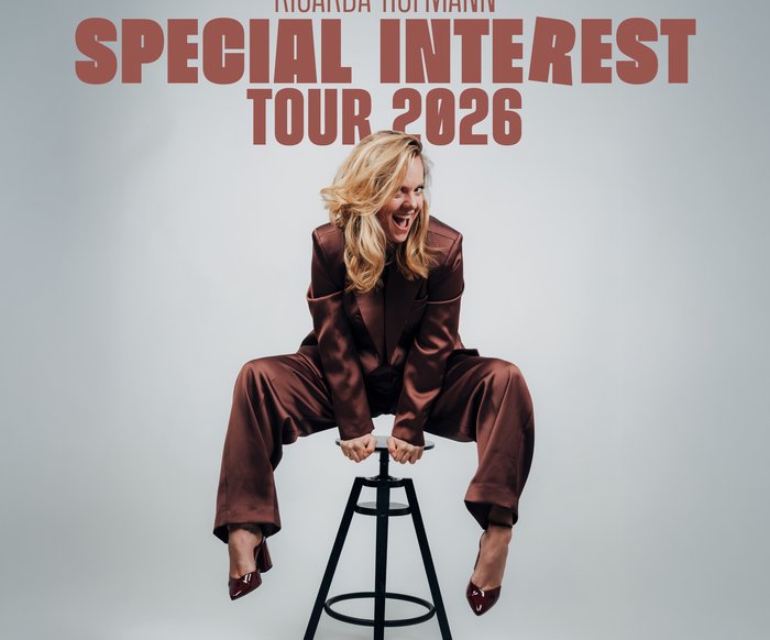 Ricardo Hoffmann in einem braunen glänzenden Jumpsuit auf einem Barhocker. Sie drückt sich mit den Händen auf der Sitzfläche hoch.