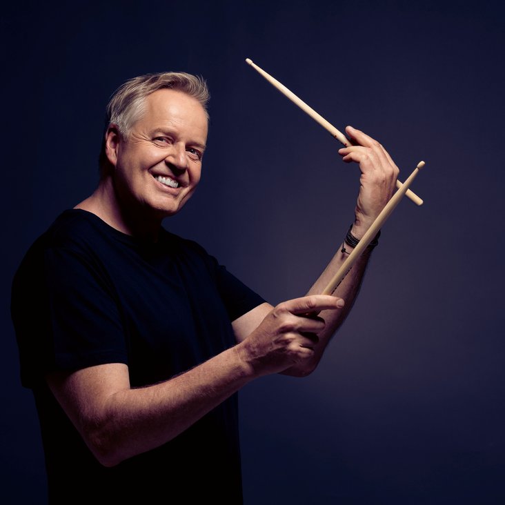 Portraitfoto Wolfgang Haffner, er trägt ein schwarzes T-Shirt, blickt lächelnd in die Kamera und hat Drum Sticks in den Händen.