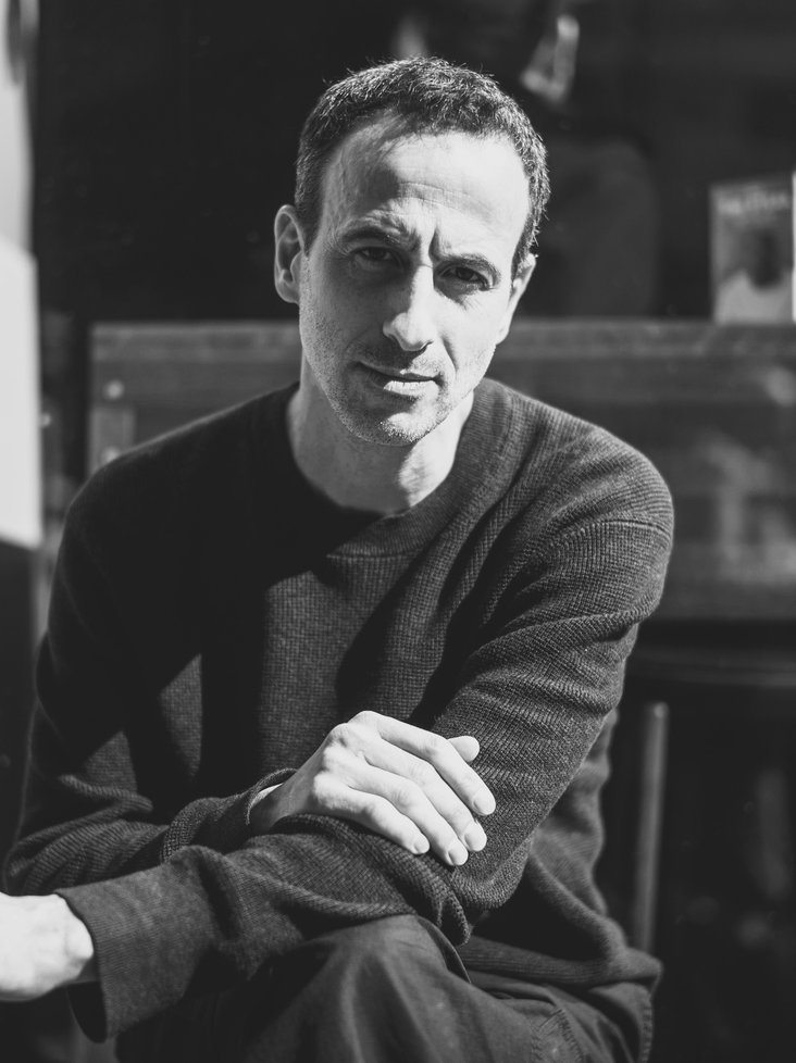 Portraitfoto von Hofesh Shechter. Das Foto ist schwarz weiß und Hofesh Shechter blickt sitzend in die Kamera.