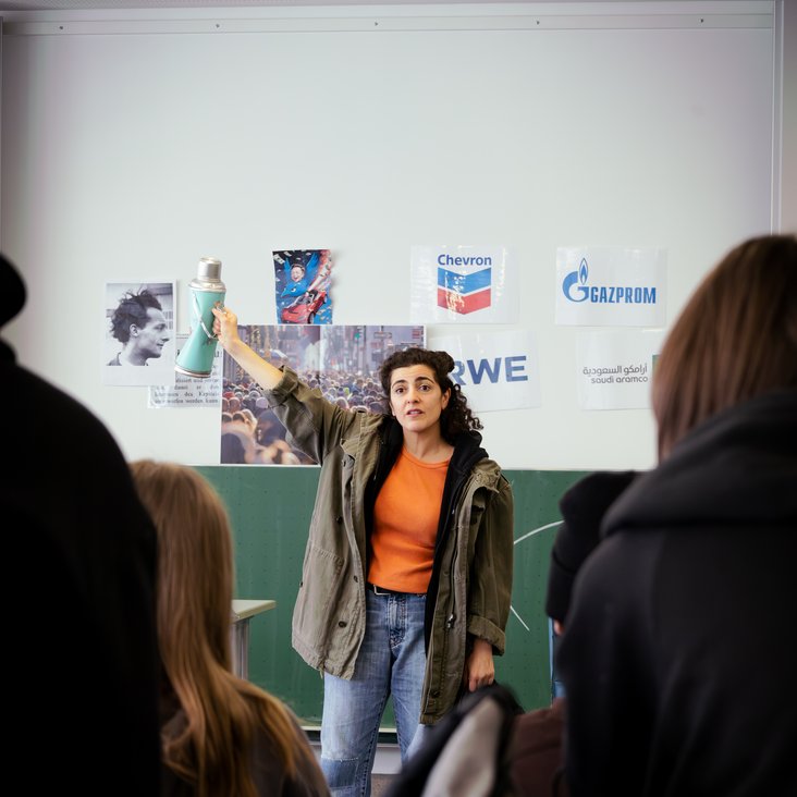 Eine Schauspielerin steht in einem Klassenzimmer vor einer Tafel. Man sieht die Schüler:innen, die ihr zusehen, von hinten.